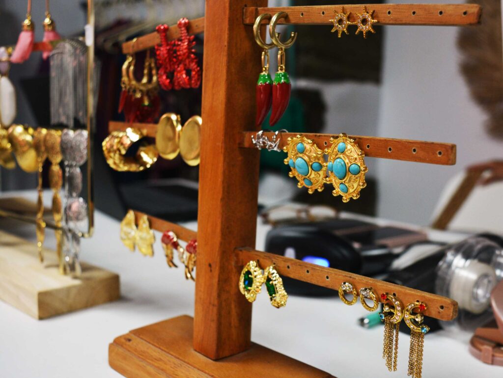 Un puesto de joyería de madera muestra pendientes de colores, incluidos diseños dorados, turquesas y rojos. Al fondo, sobre una superficie blanca, se ve otro soporte con más pendientes.