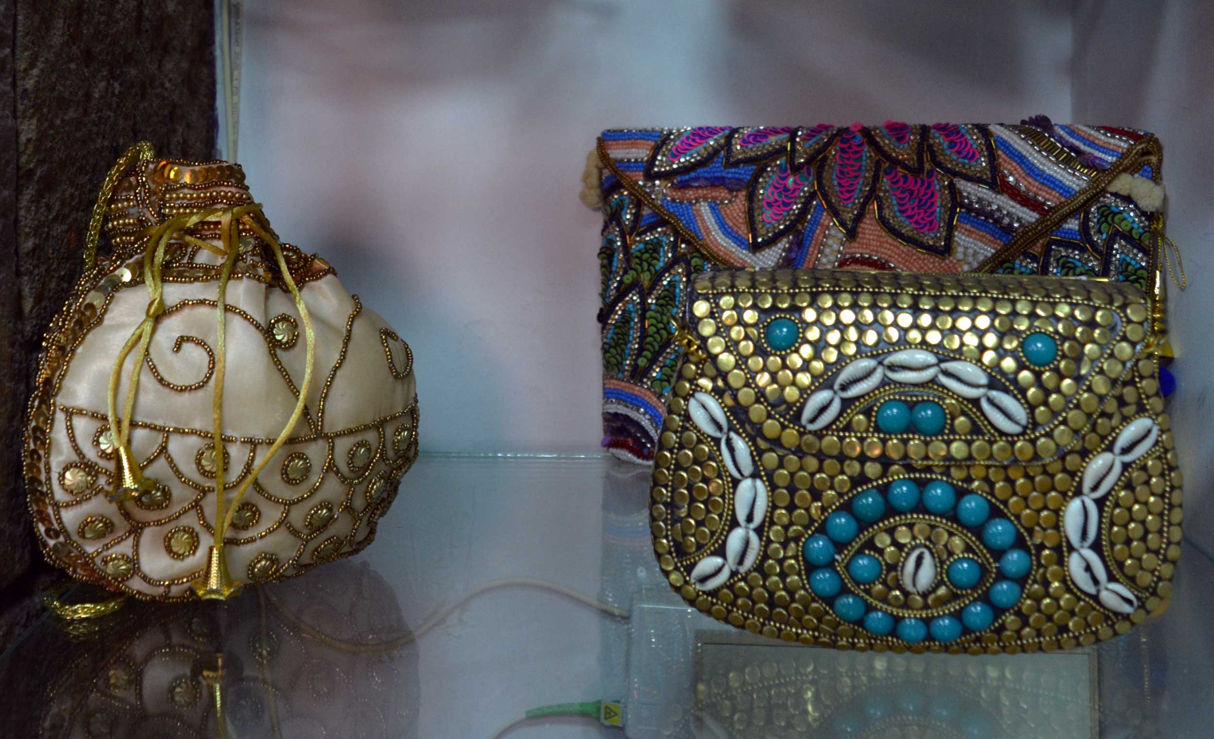 Se exponen tres bolsos decorativos: uno blanco con remolinos y abalorios dorados, otro rectangular con bordados de colores y otro dorado con abalorios turquesa, conchas de cauri y dibujos intrincados.