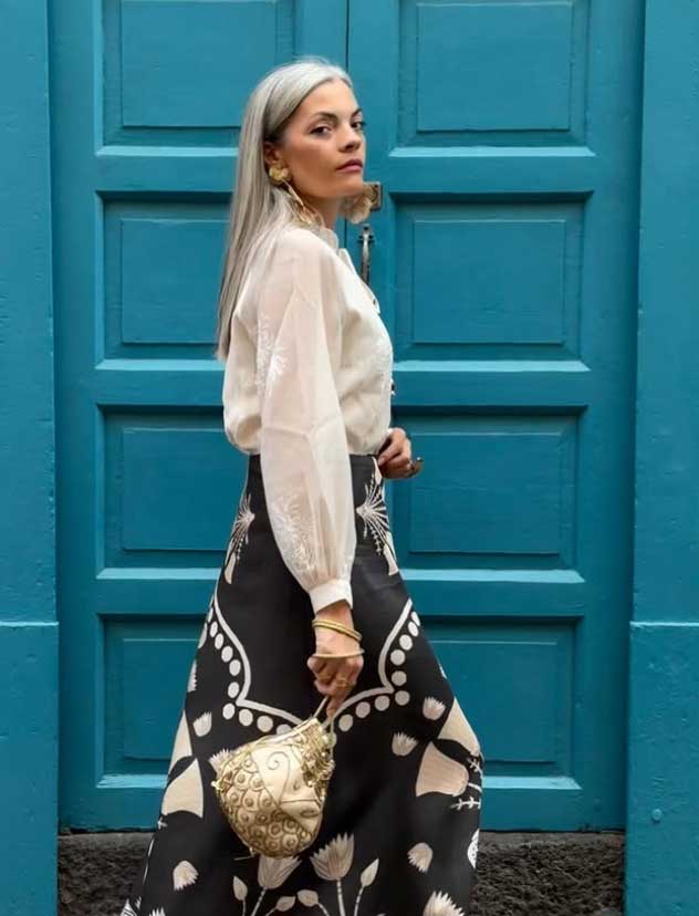 Una mujer de pelo largo y canoso, de pie delante de una puerta azul, lleva una blusa blanca transparente, una falda negra vaporosa con motivos florales blancos, pendientes de oro, pulseras y un bolso de mano con textura dorada.
