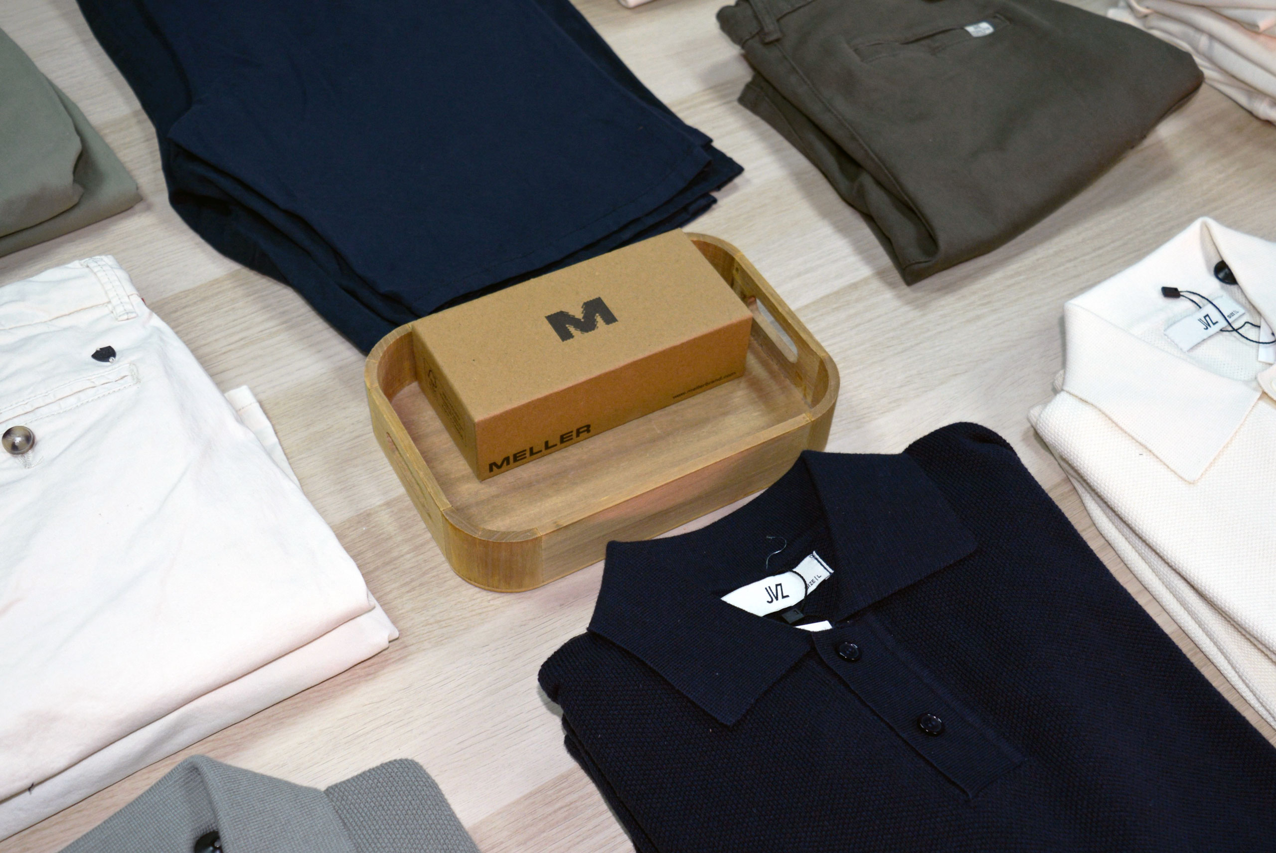 Un muestrario ordenado de ropa doblada, que incluye prendas de color azul marino, blanco, beige y oliva, con una caja marrón con la etiqueta MELLER colocada en una bandeja de madera en el centro.