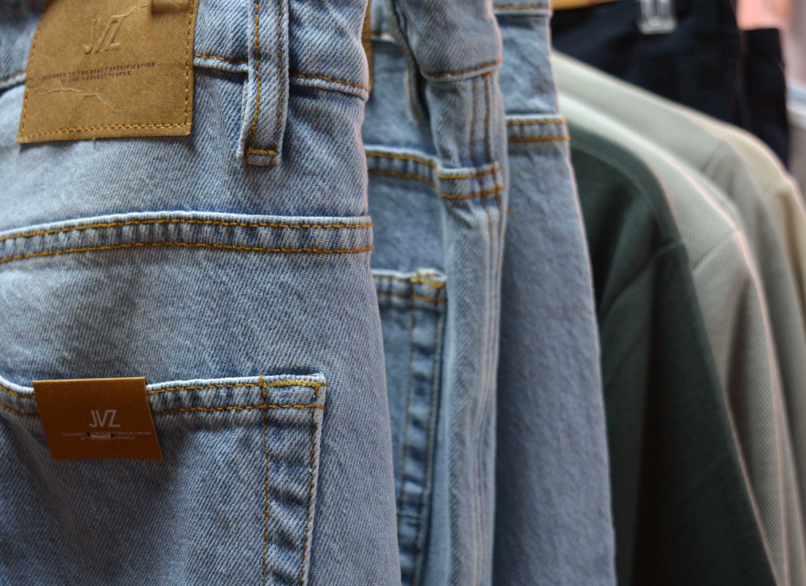 Primer plano de unos vaqueros azules colgados en un perchero, mostrando los bolsillos traseros y los parches de cuero marrón de la marca, con camisas difuminadas en colores verde, negro y gris al fondo.