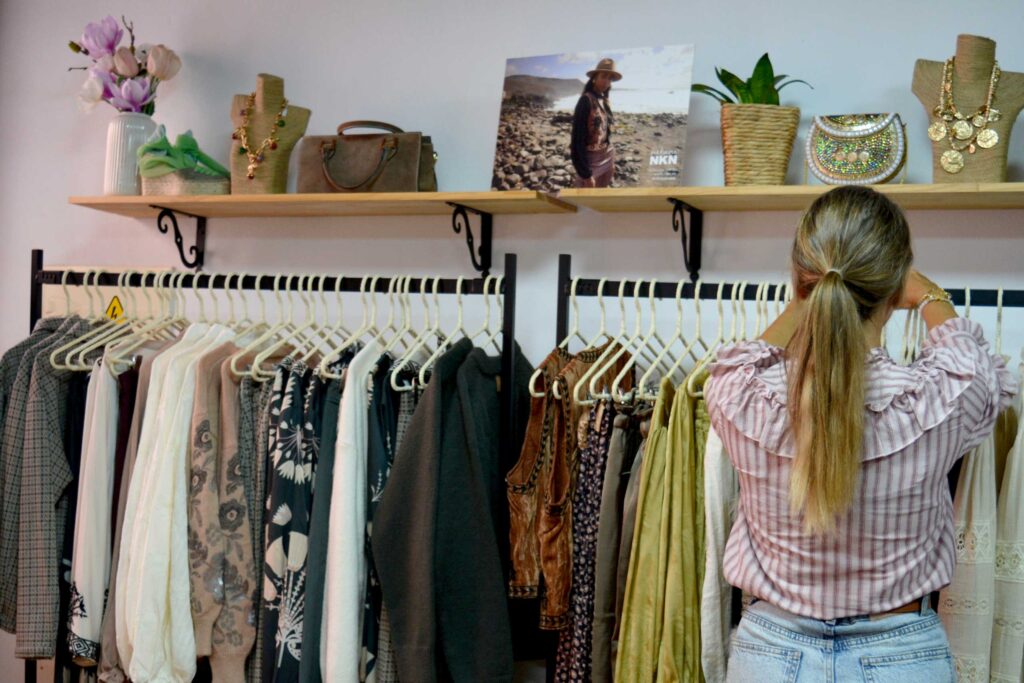 Una mujer con coleta ojea ropa en percheros bajo estanterías con bolsos, joyas y una foto decorativa en una tienda boutique.
