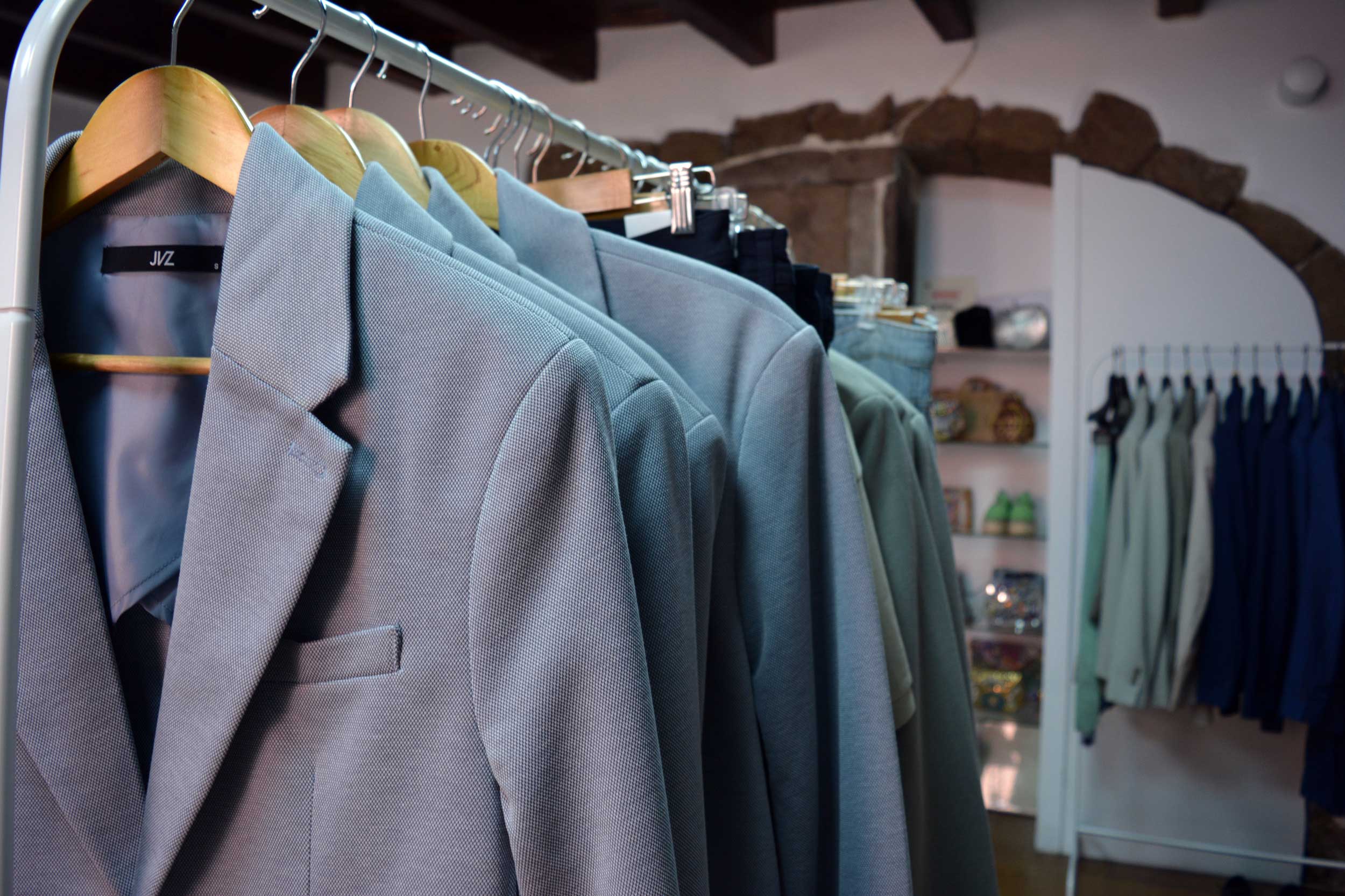 Varias americanas gris claro cuelgan de perchas de madera en un perchero de ropa en una boutique. Al fondo, hay estanterías con zapatos y más ropa colgada en un perchero.